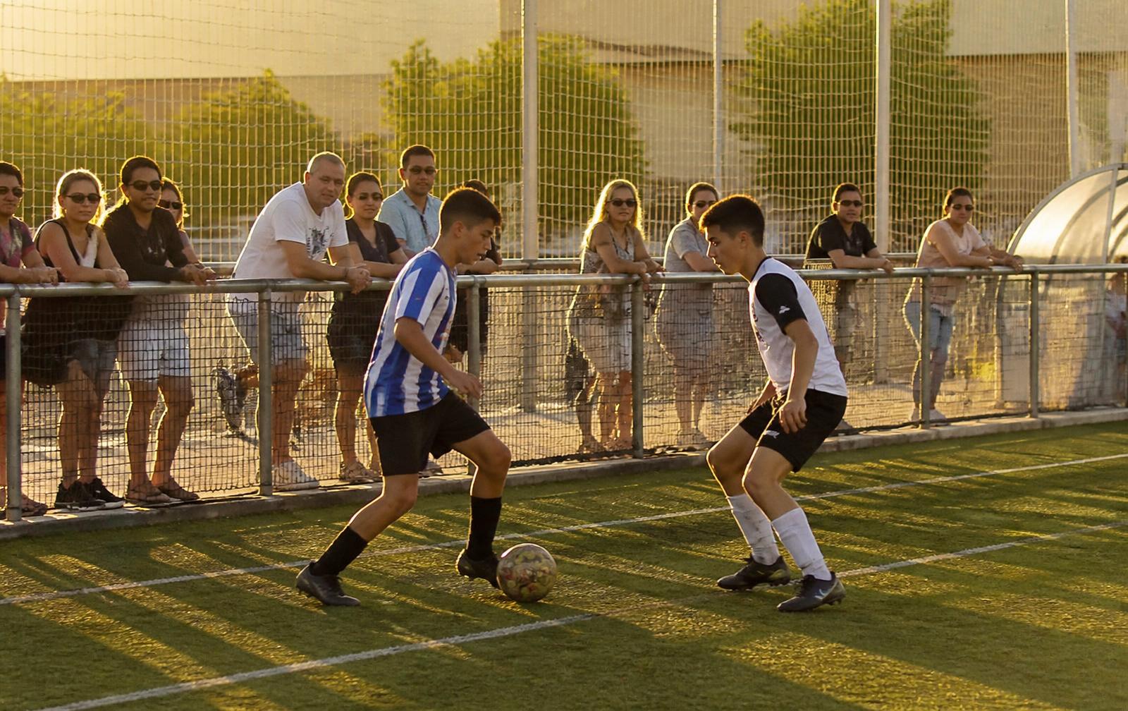 Partido de fútbol amateur en un campo de barrio con familias animando desde la valla